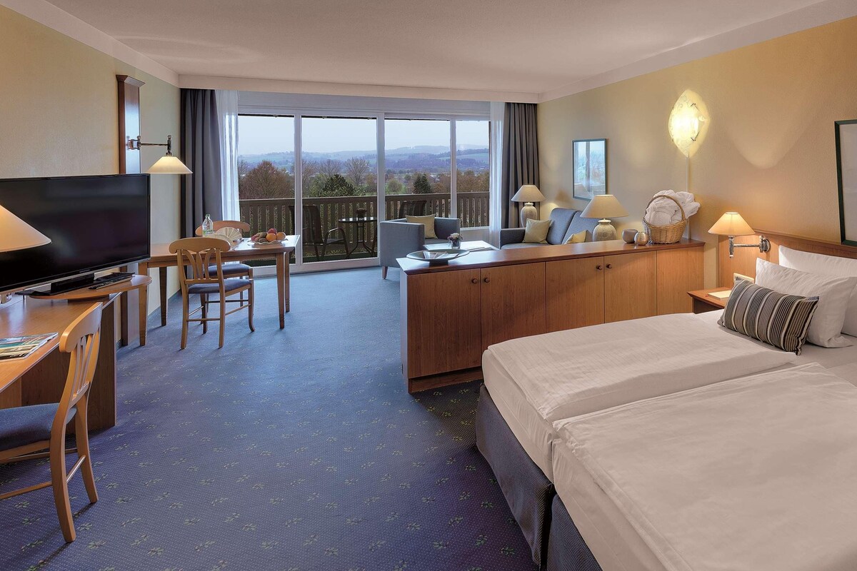 Habitación de hotel con una cama grande, un escritorio con televisor, un balcón con vistas y un comedor.