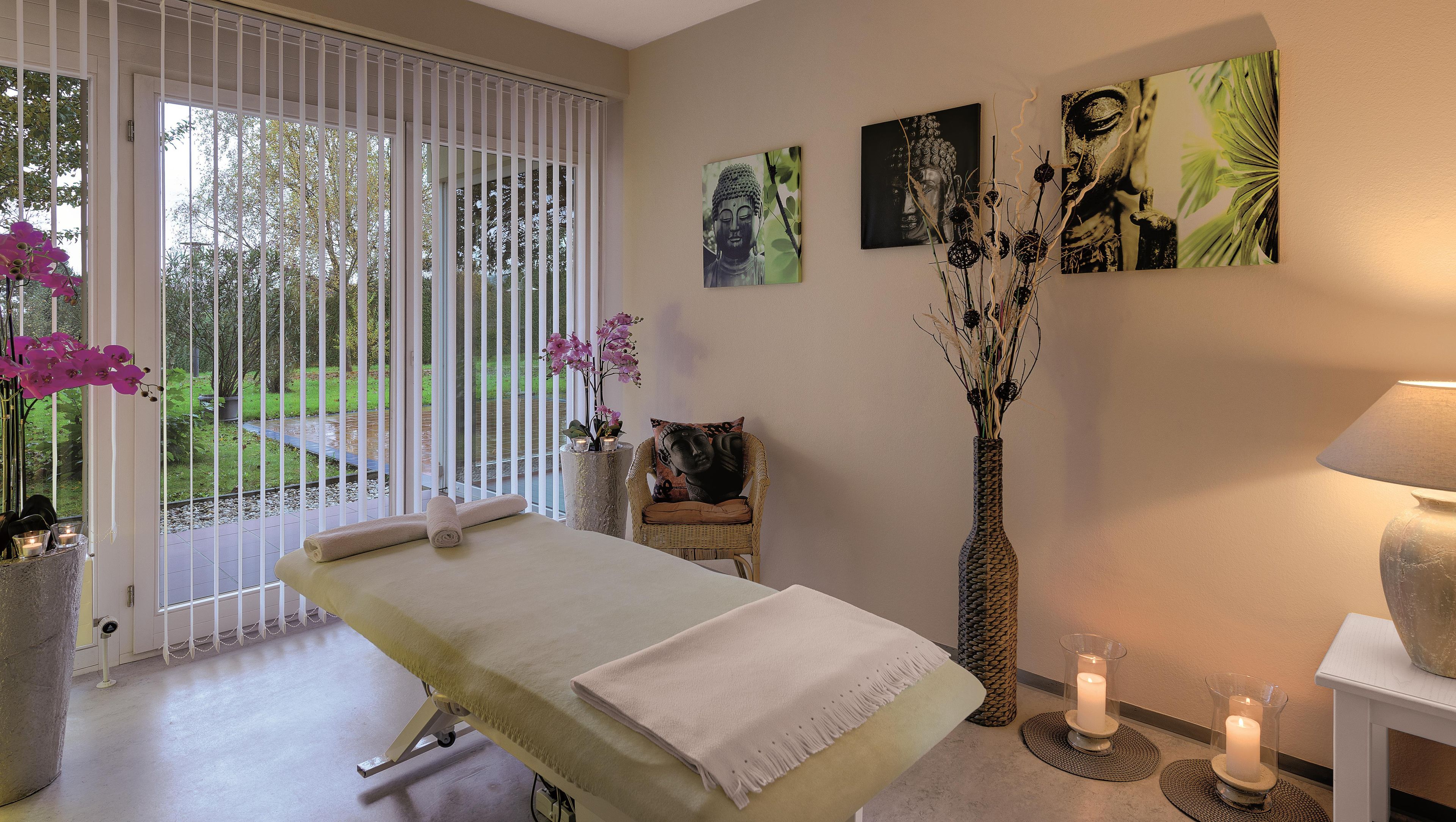 A spa room with a massage table, a chair, a vase with flowers, and wall art.