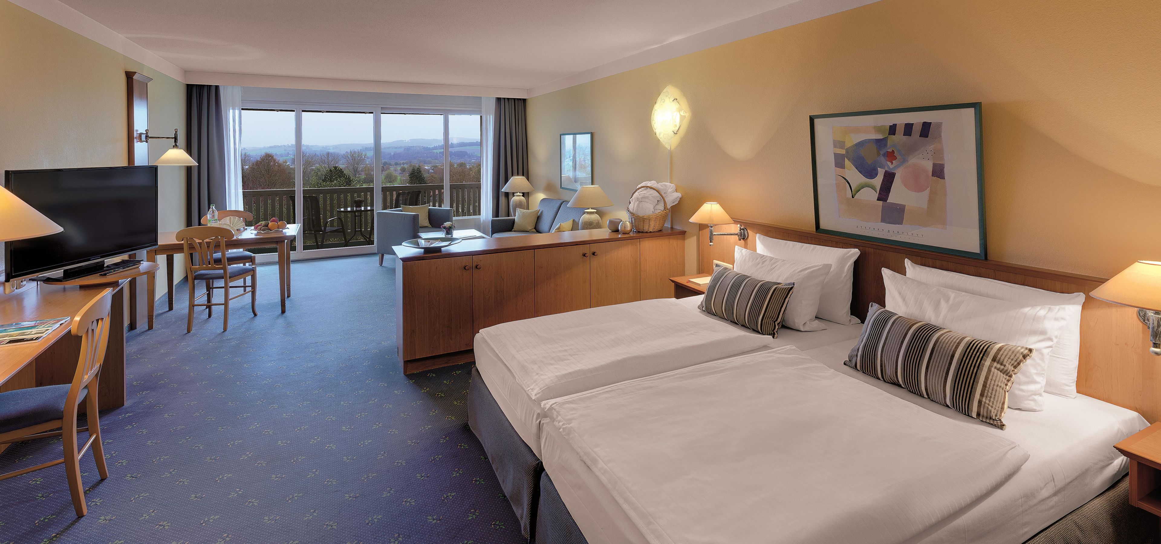 Ein Hotelzimmer mit einem großen Bett, zwei Nachttischen, einem Schreibtisch mit Fernseher, einem Essbereich mit Stühlen und Blick ins Freie.