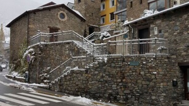 A stone building with a staircase leading to a balcony, a yellow building with a large window, and a road with snow on the sides. - Hotel Comapedrosa (Arinsal)