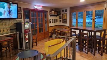 A bar area with a television displaying a sports event, a Budweiser sign, a biscuit sign, a vending machine, and a view of the outside through large windows.