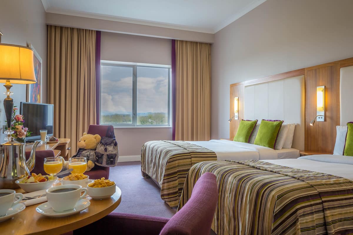 Habitación de hotel con dos camas, zona de comedor con desayuno, ventana grande y vistas al exterior.