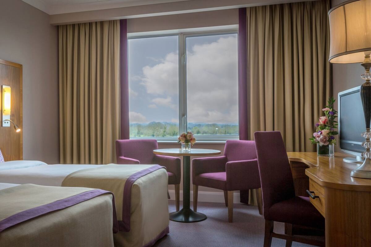 Habitación de hotel con dos camas, un escritorio con computadora, una silla, una mesita y una ventana con vista al exterior.