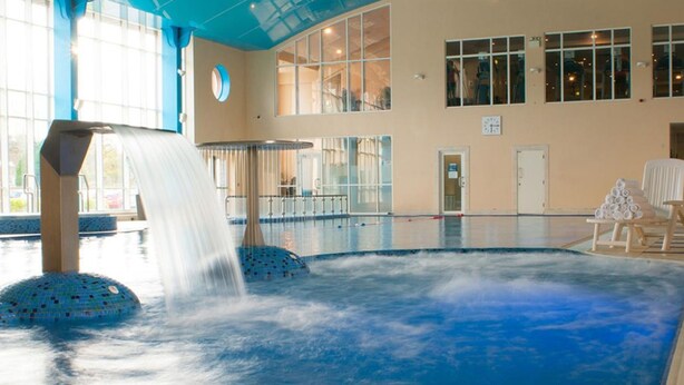 Una gran piscina interior con una cascada, rodeada por una terraza con baldosas y una pared de vidrio.