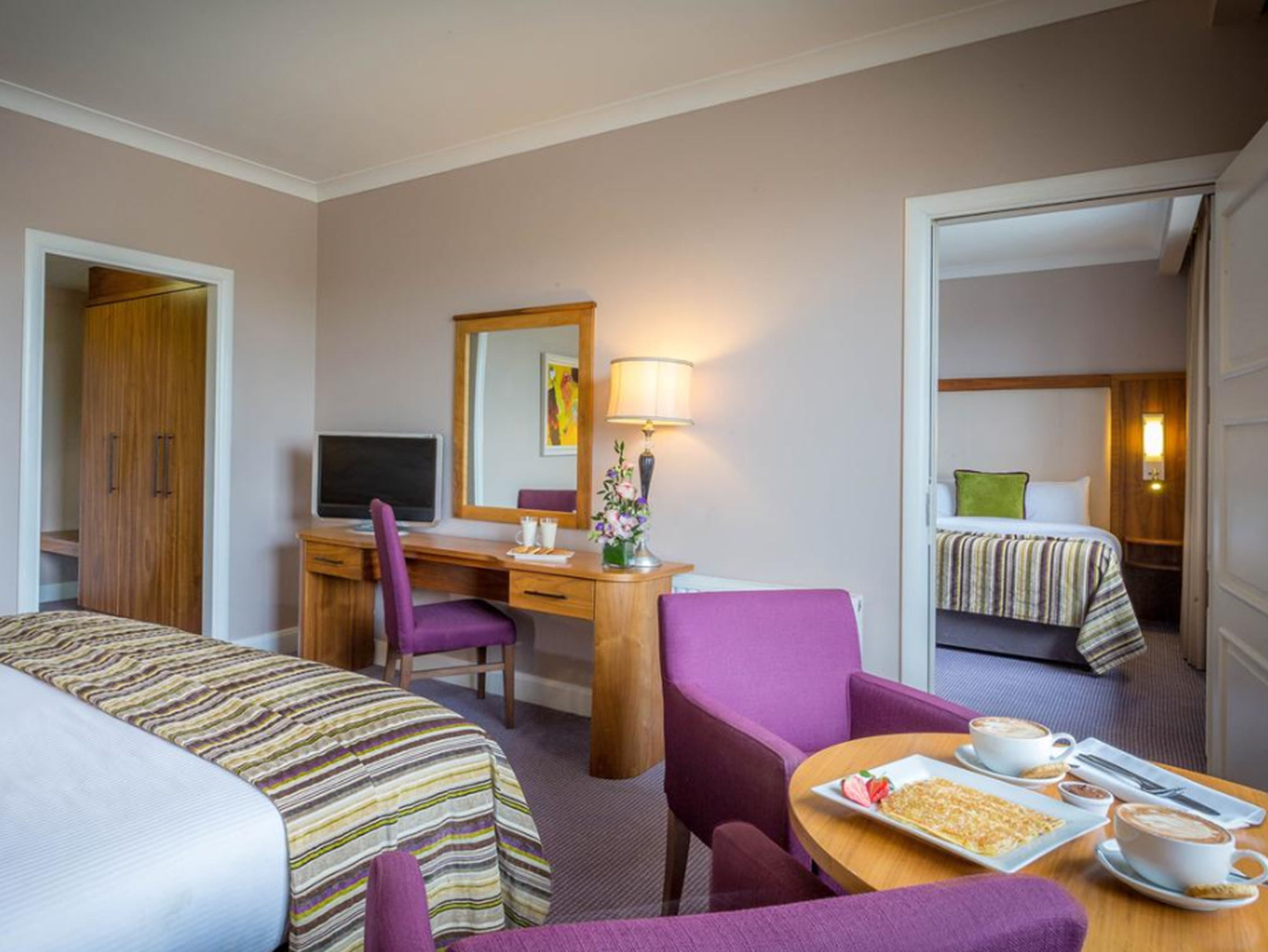 A hotel room with two beds, a desk with a computer, a chair, a table with breakfast, and a bathroom visible through an open door.