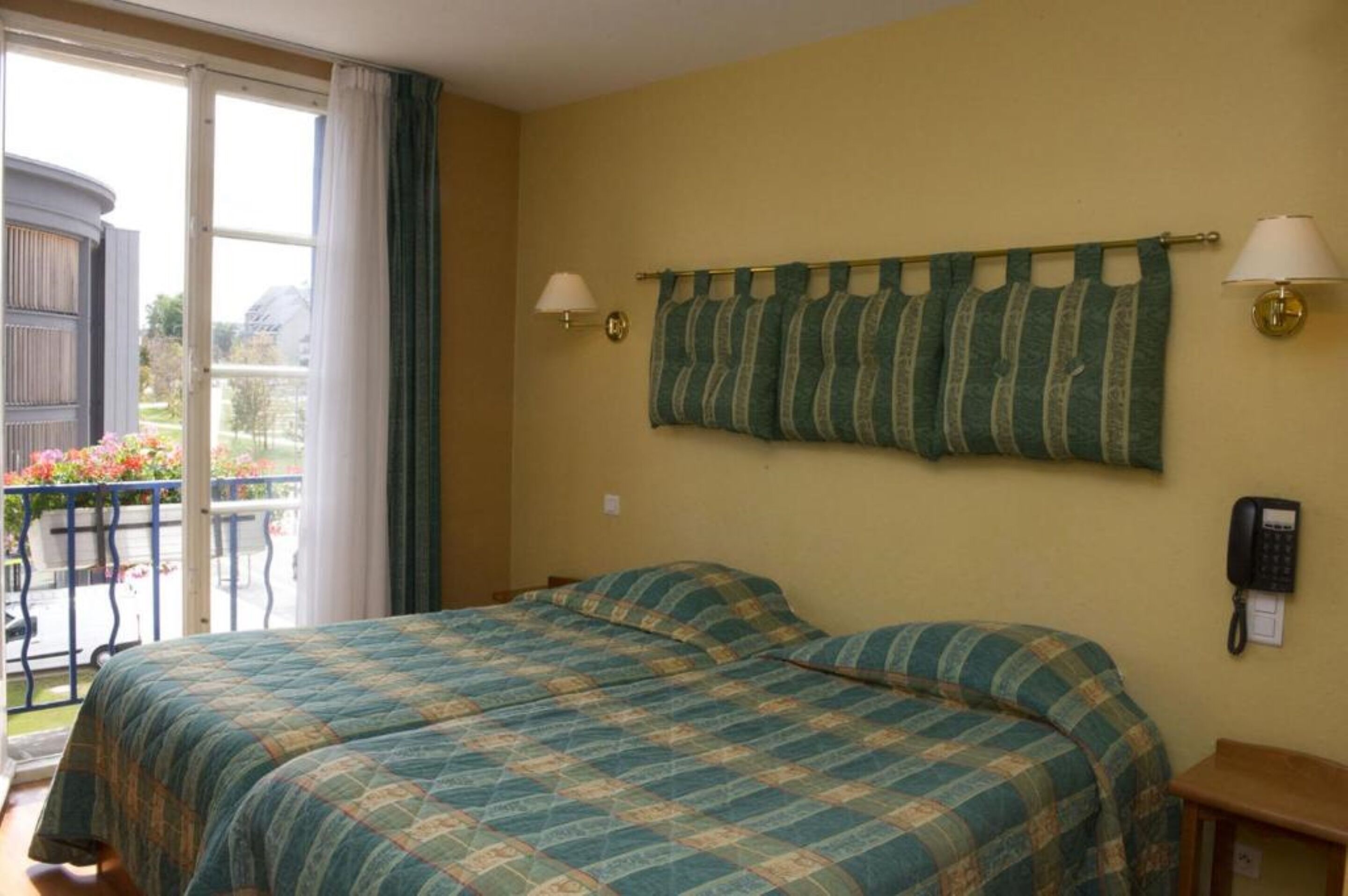 Habitación de hotel con cama, una mesita, teléfono y vistas a un balcón lleno de flores.