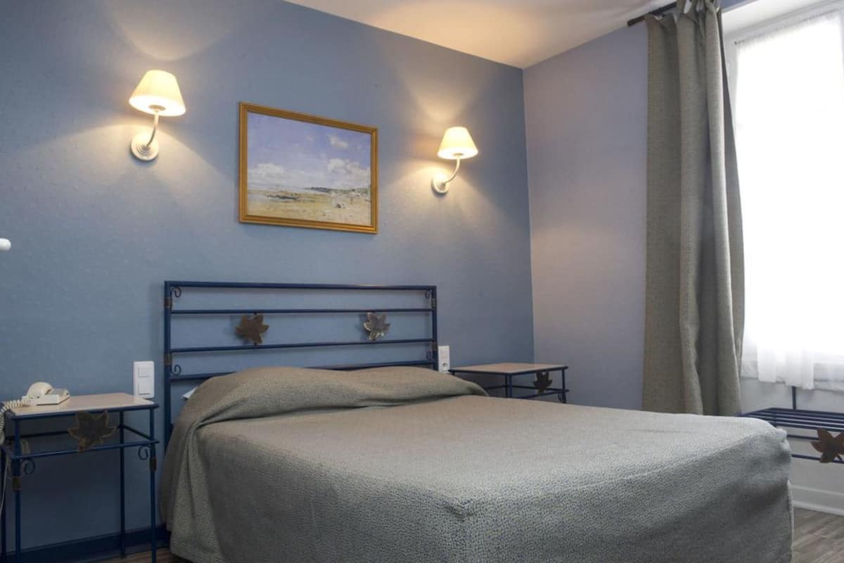 A hotel room with a bed, two bedside tables, a lamp, a painting, and a window with curtains.