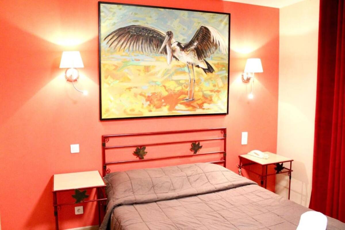 A room with a red bed, two bedside tables with lamps, and a painting of a bird on the wall.