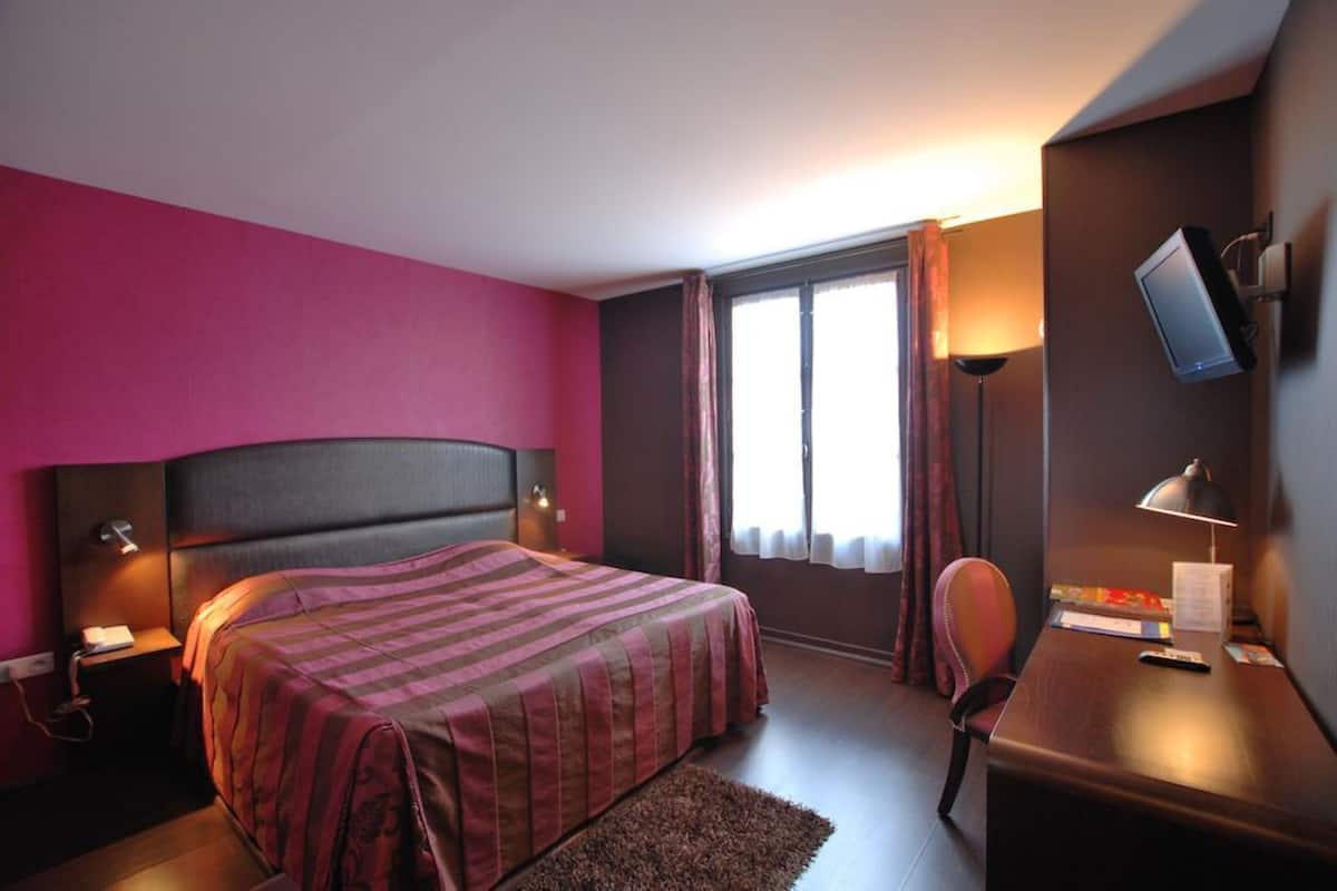 A hotel room with a bed, bedside tables, a desk, a chair, a TV, and a window with curtains.