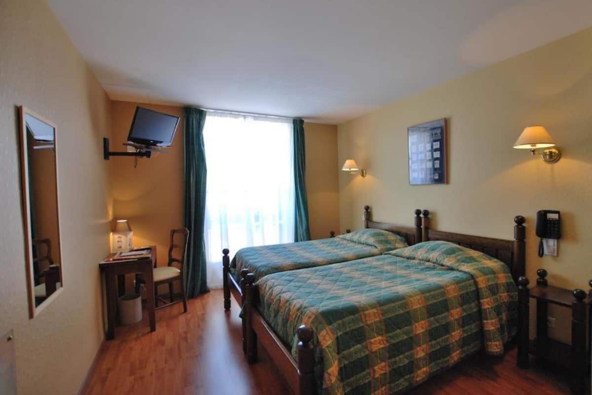 A hotel room with two beds, a TV mounted on the wall, a desk with a chair, and a window with curtains.