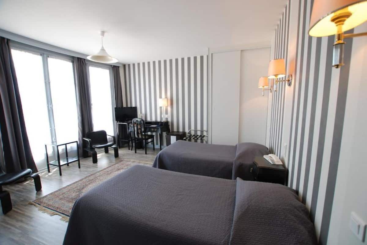 A hotel room with two beds, a desk with a chair, a television, and a large window with curtains.