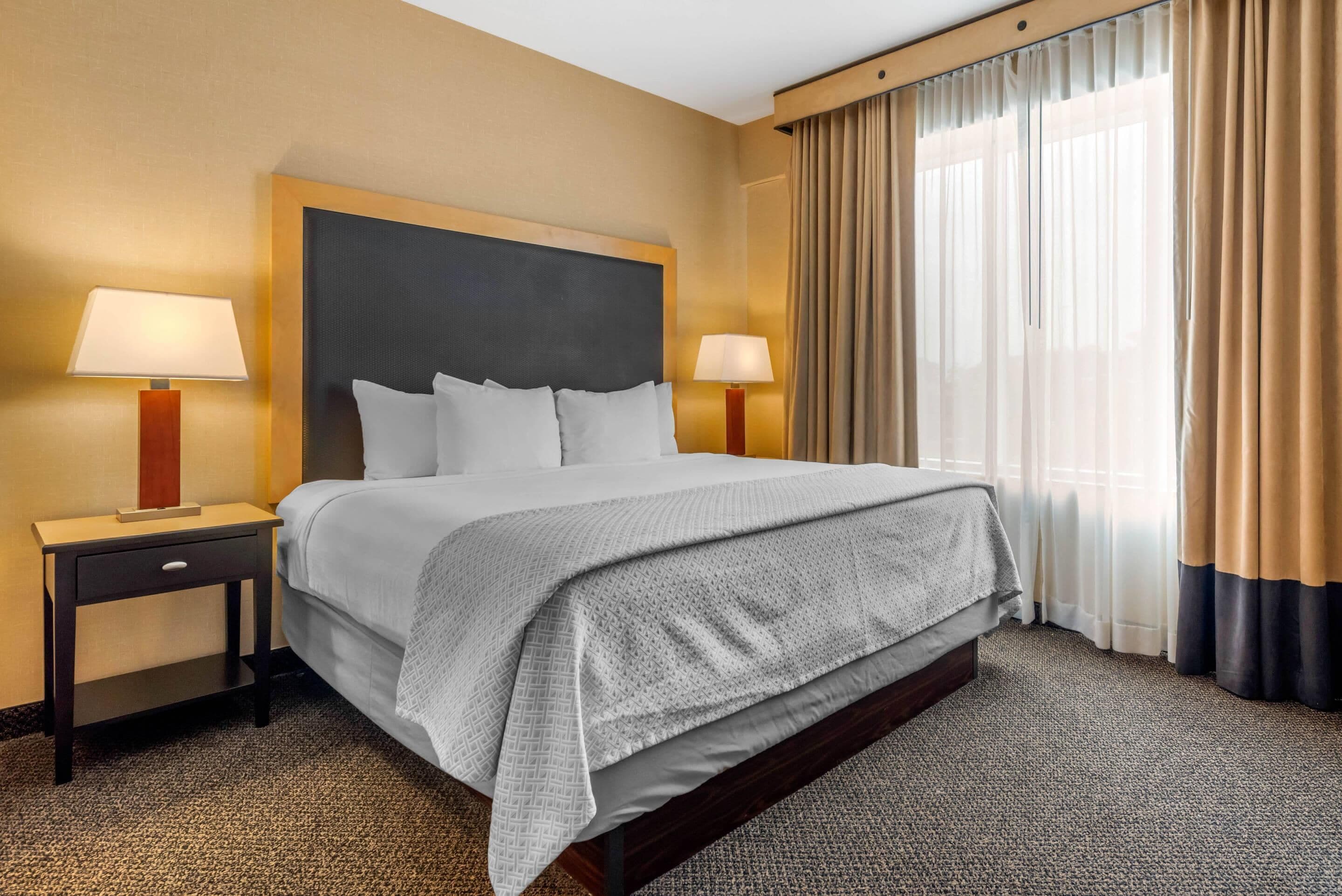 A hotel room with a large bed, two bedside tables with lamps, a headboard, and a window with curtains.