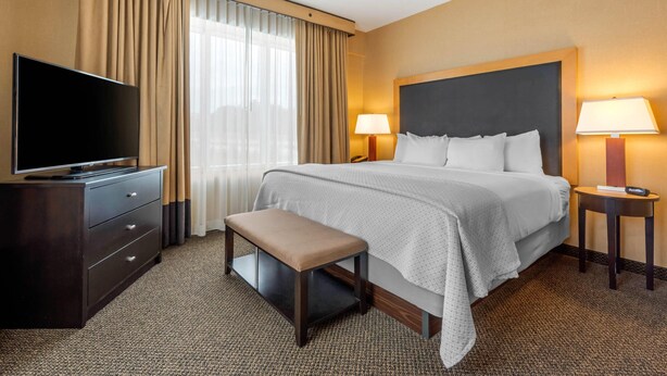 Una habitación de hotel con una cama, televisión, una banca, dos lámparas de noche y un ventanal con cortinas.