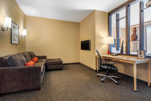 A hotel room with a gray sofa, a small ottoman, a desk with a chair, a flat-screen TV, and a large window with a vase.
