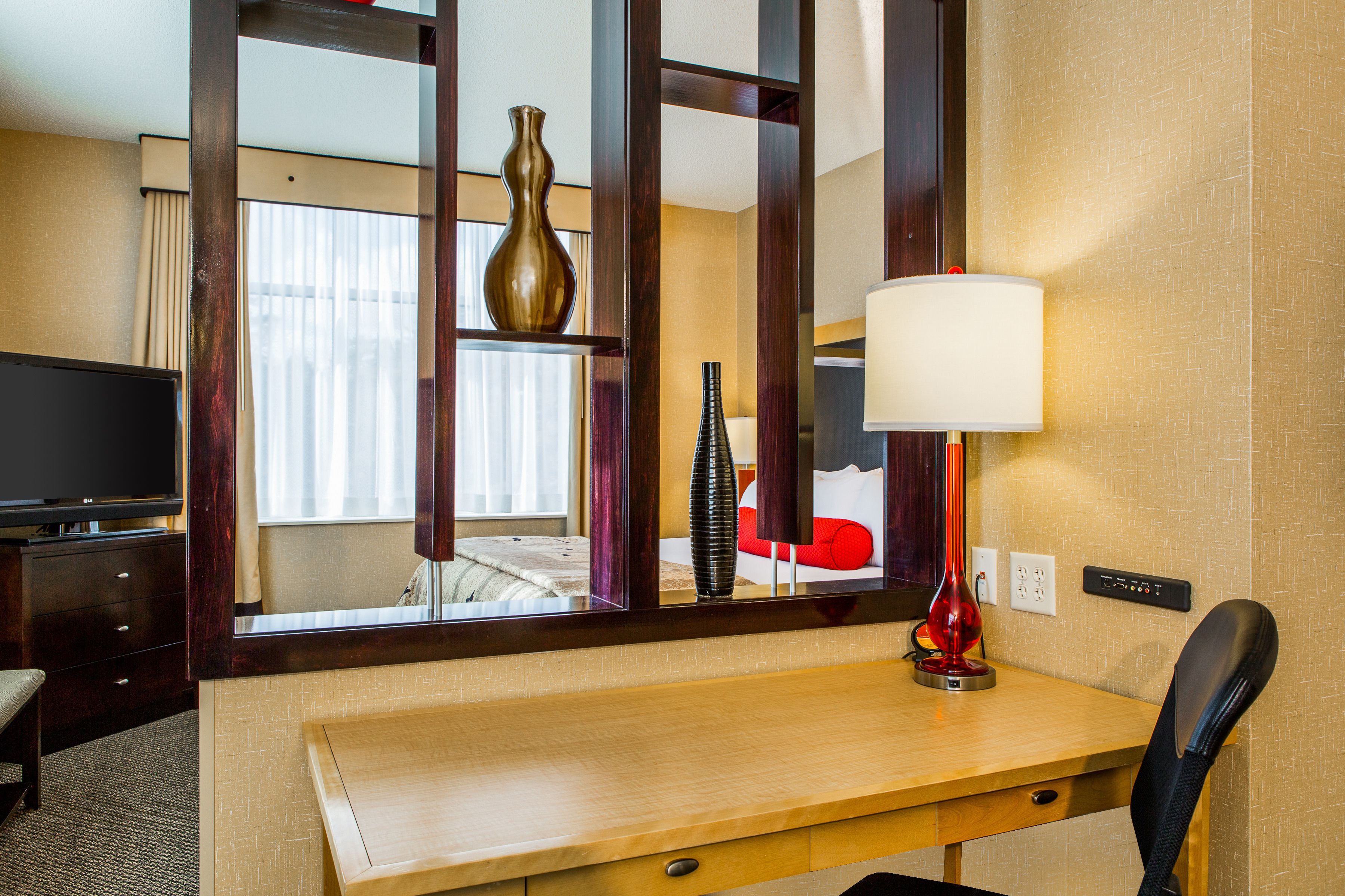 A hotel room with a wooden desk, a television, a red lamp, and a large mirror reflecting the room.