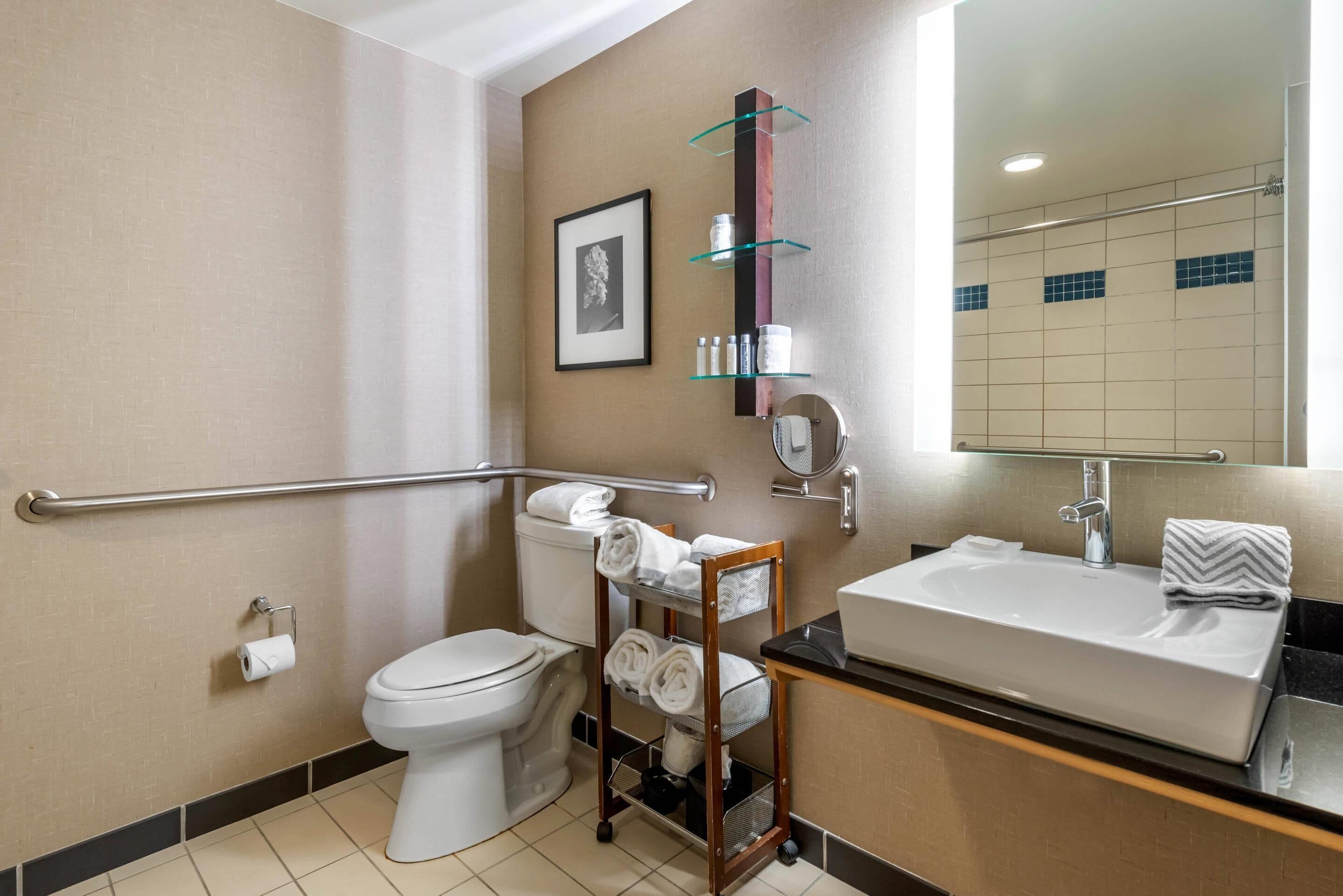 Un baño moderno con inodoro, lavamanos, espejo, toallero y barra de apoyo.