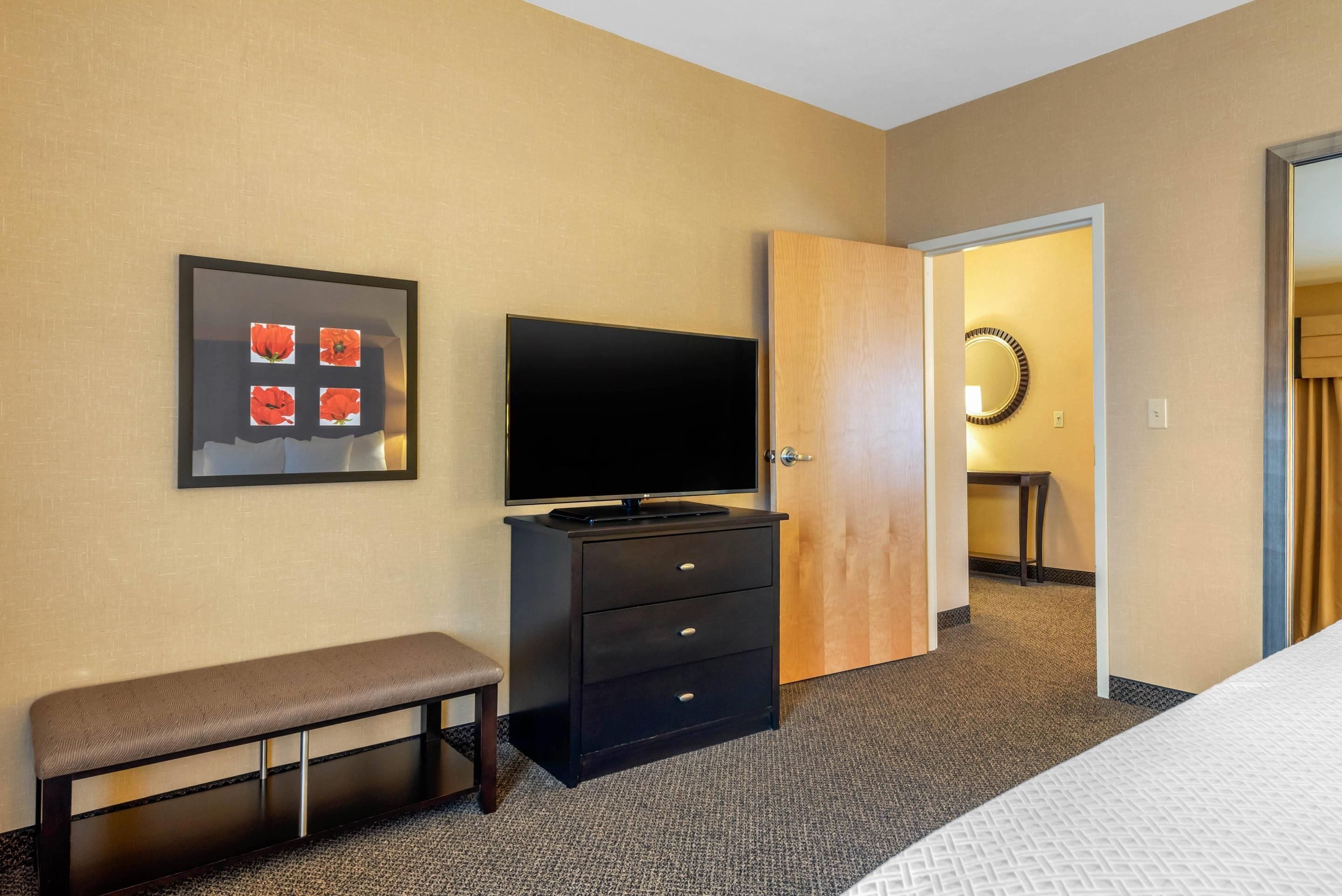 A hotel room with a bed, a television on a dresser, a bench, a door leading to a bathroom, and a framed picture on the wall.