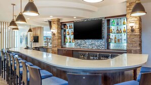 A modern bar area with a curved counter, multiple bar stools, and a variety of liquor bottles. - Cambria Hotel Akron - Canton Airport (Uniontown)