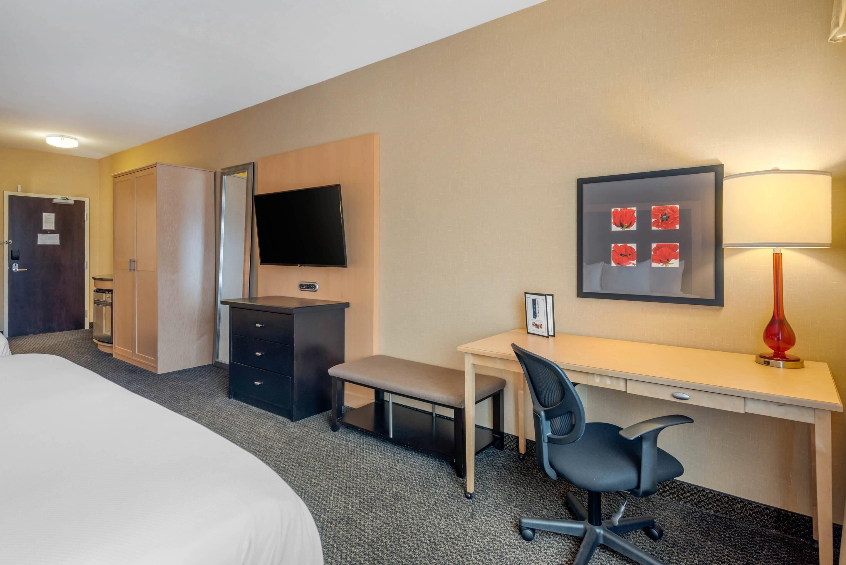 Una habitación de hotel con cama, escritorio, silla, televisor, armario y lámpara.