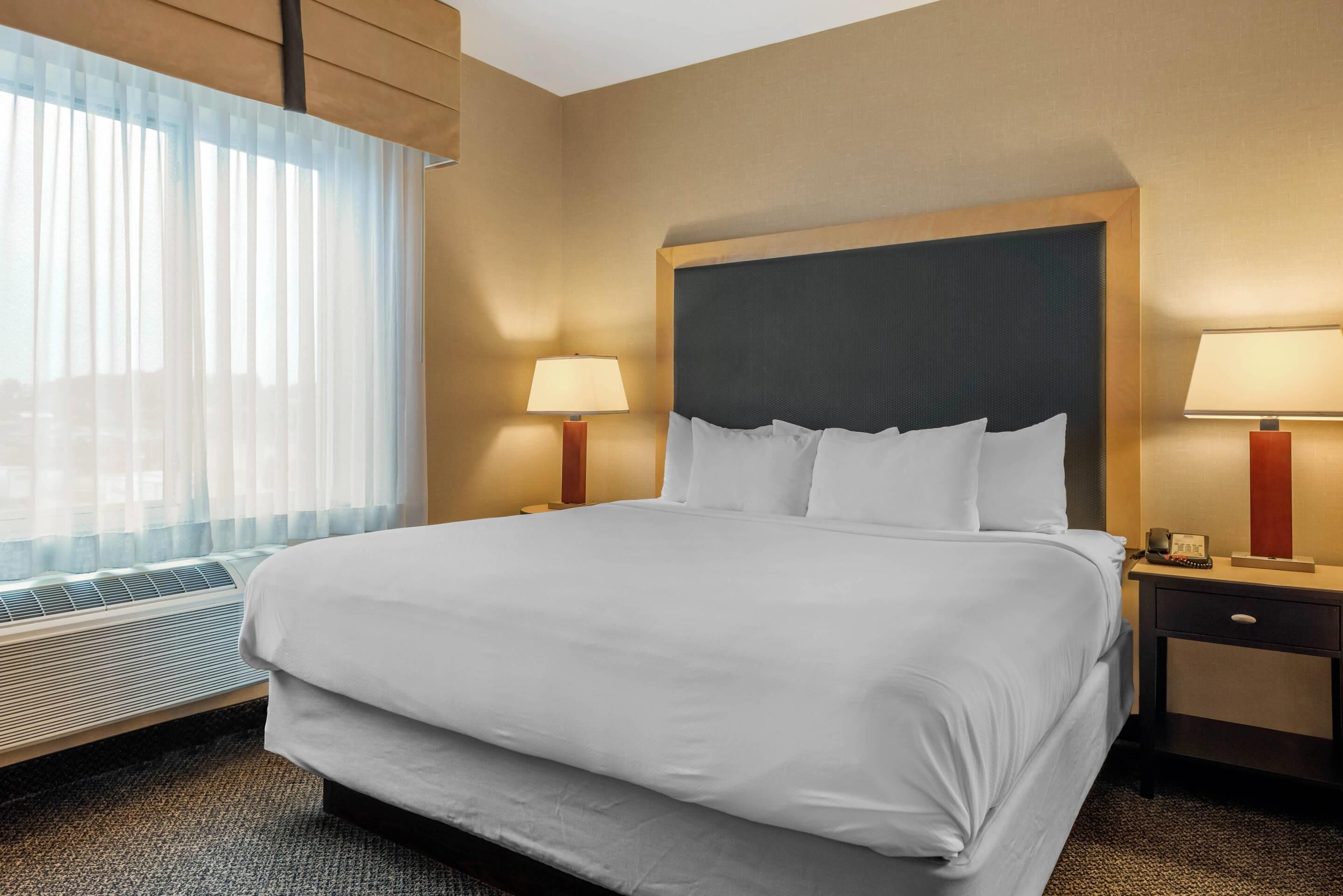 Hotelkamer met een groot bed, twee bedlampen, een verwarming en een raam met dunne gordijnen.
