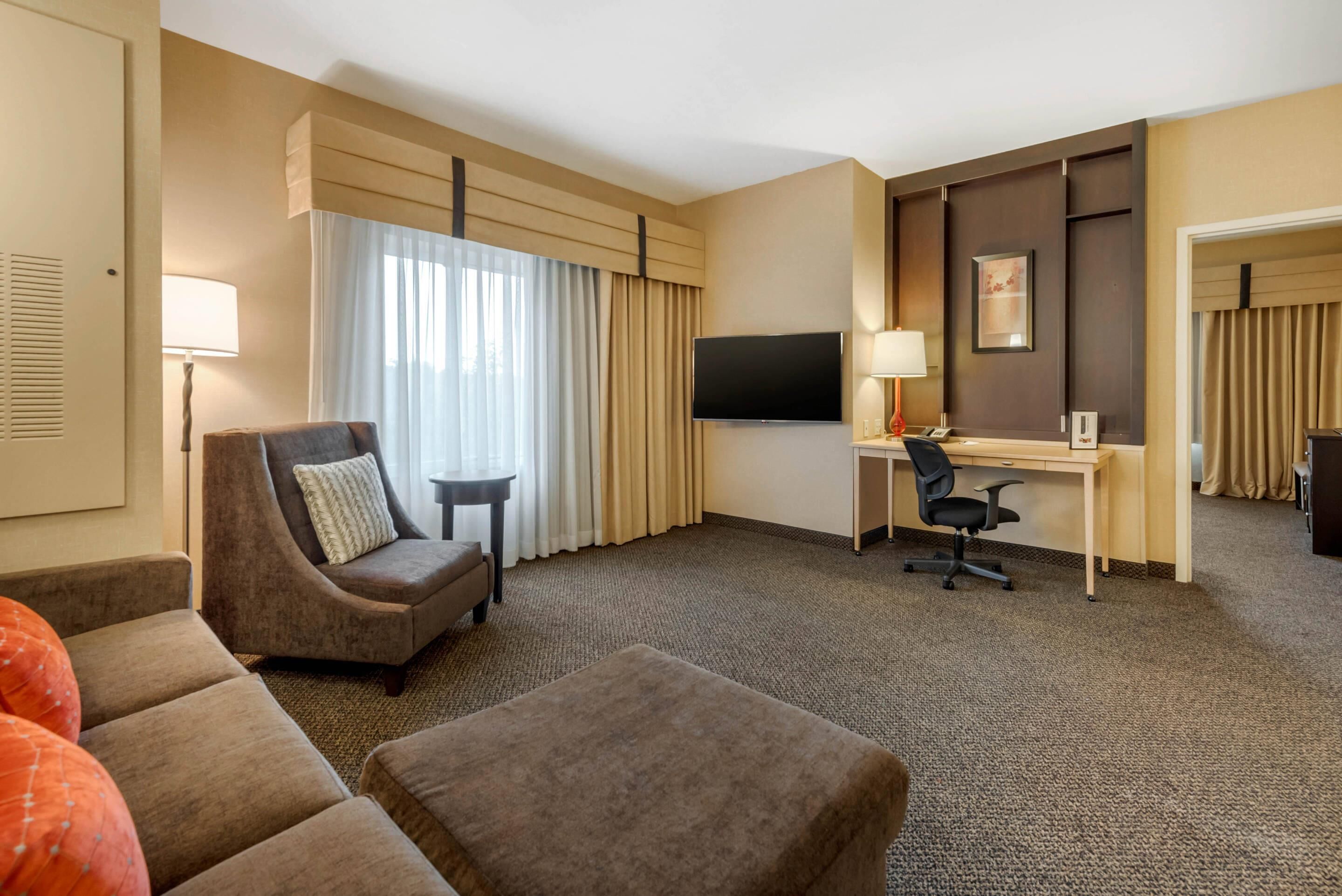 Una habitación de hotel con sofá, escritorio, televisión y ventana con cortinas.