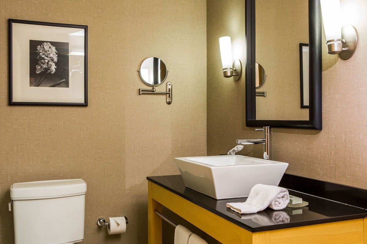 Une salle de bain avec un lavabo blanc, des toilettes, un tableau encadré au mur et un miroir avec des lumières.