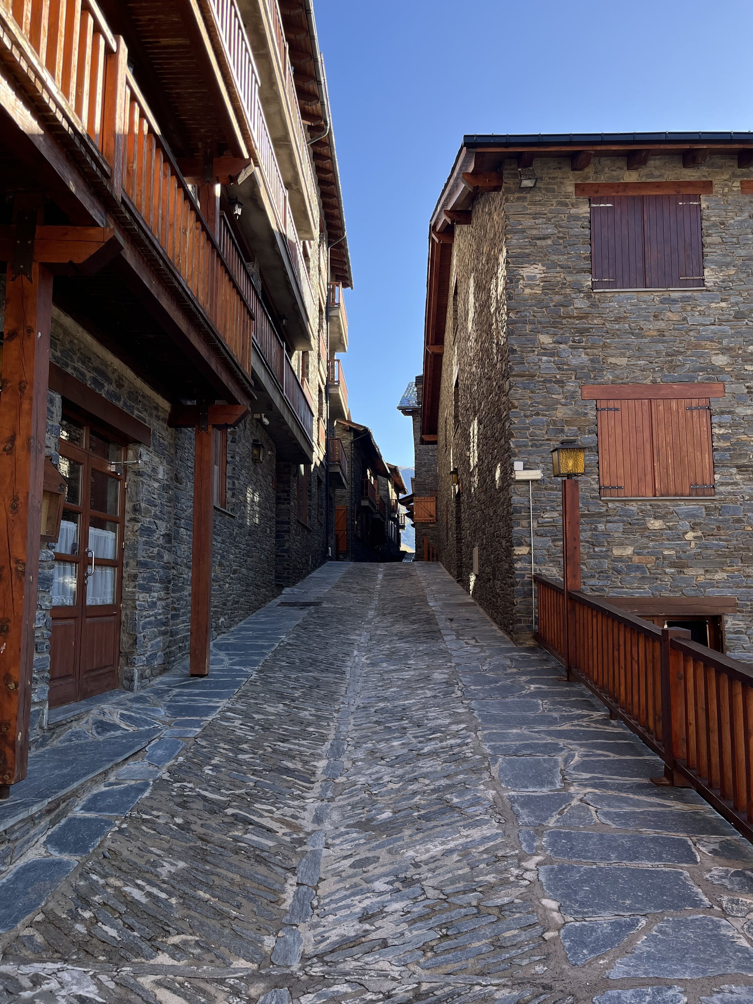 A narrow cobblestone street flanked by stone buildings with wooden balconies and shutters.