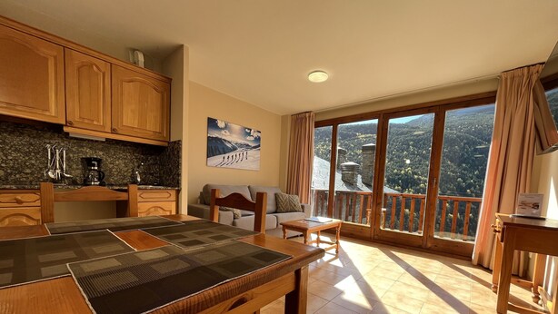 A cozy dining area with a wooden table and chairs, a kitchen with granite countertops, and a large window offering a scenic mountain view. - Wuau! Apartaments Font del Poble (Ransol)