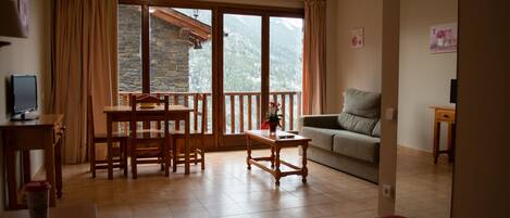 A cozy living room with a wooden dining table, a sofa, and a balcony with a mountain view.