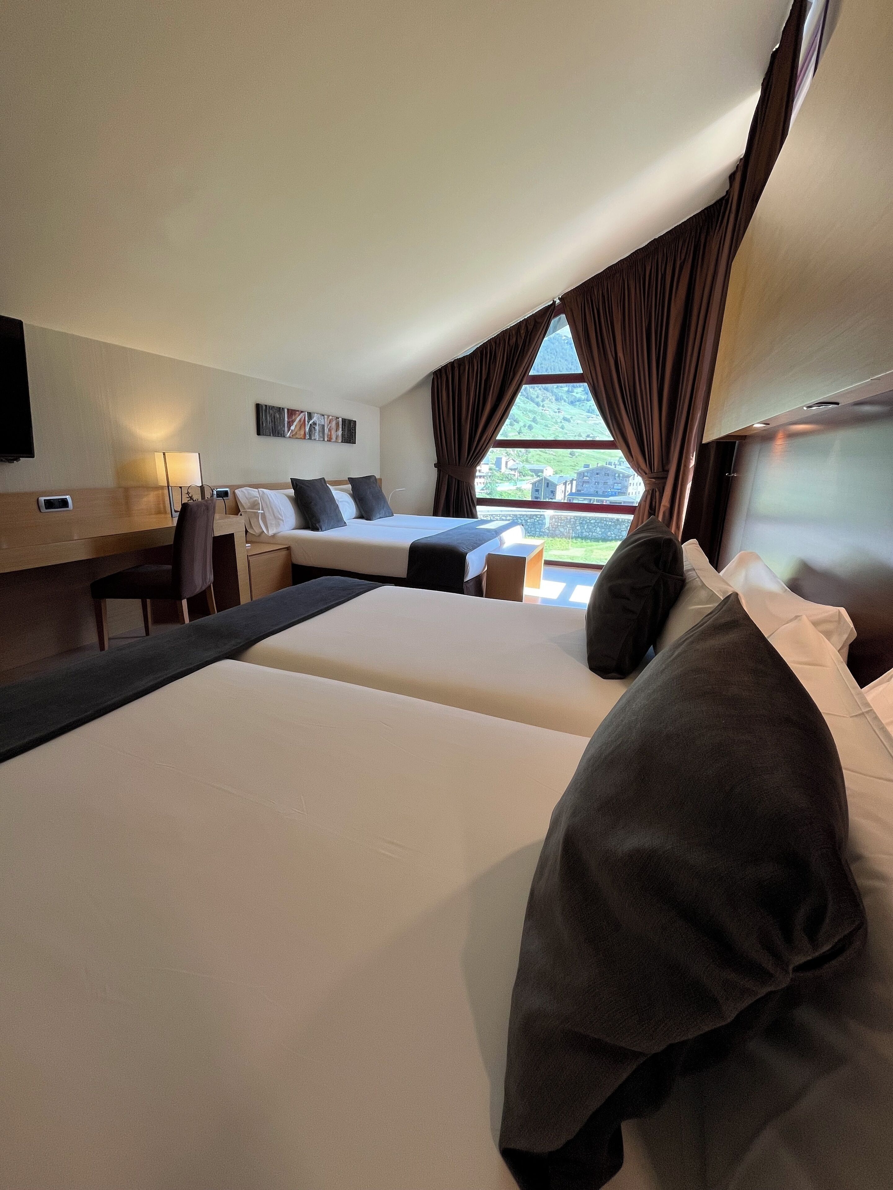 A hotel room with two beds, a desk, a chair, a TV, and a window with curtains.
