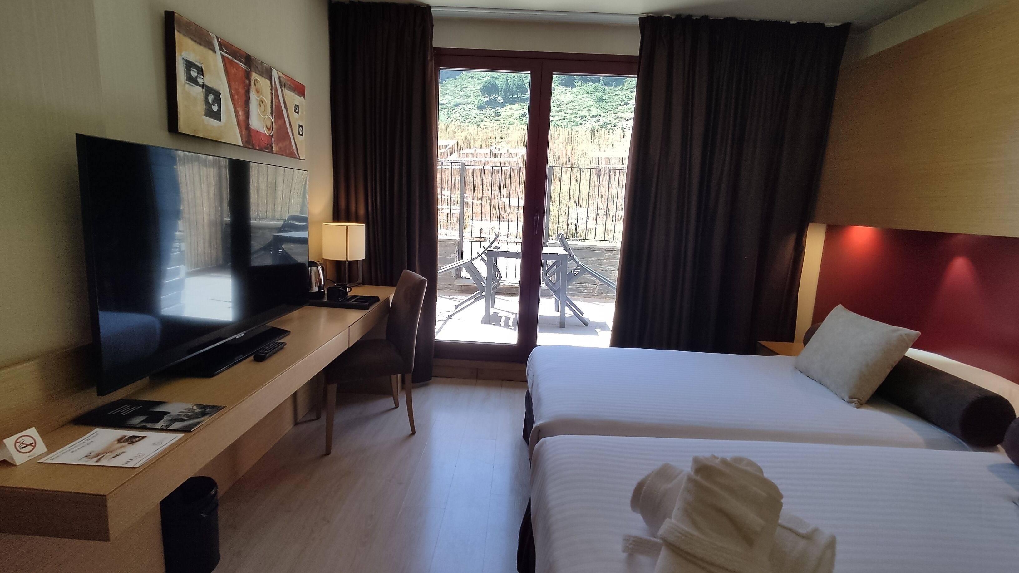 A hotel room with a TV, desk, chair, two beds, and a balcony with outdoor furniture.