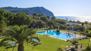 Ein Swimmingpool mit Liegestühlen und Sonnenschirmen, umgeben von Grünflächen und einem Berg im Hintergrund.