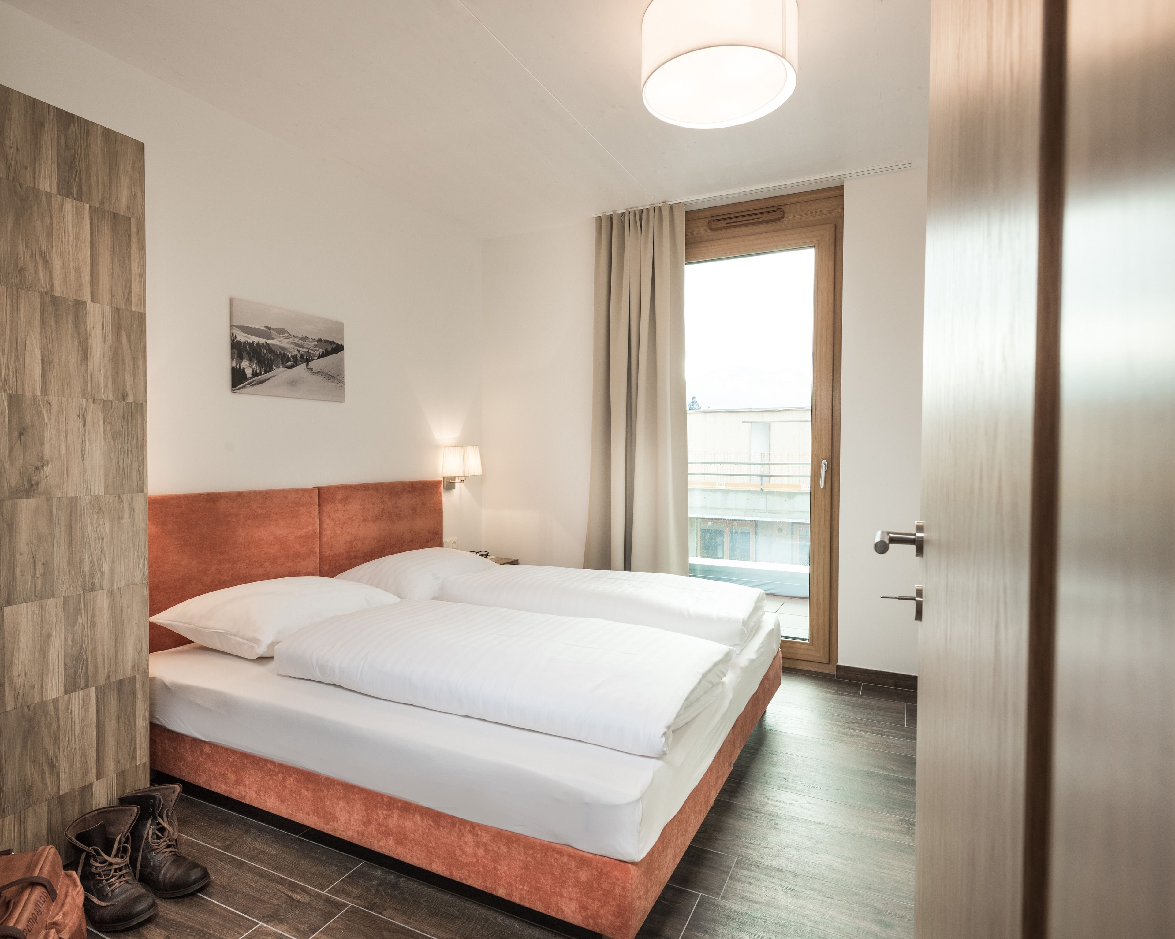 Une chambre d’hôtel avec un lit double, une tête de lit en bois, une table de chevet avec une lampe, une paire de bottes et une vue sur un immeuble par la fenêtre.