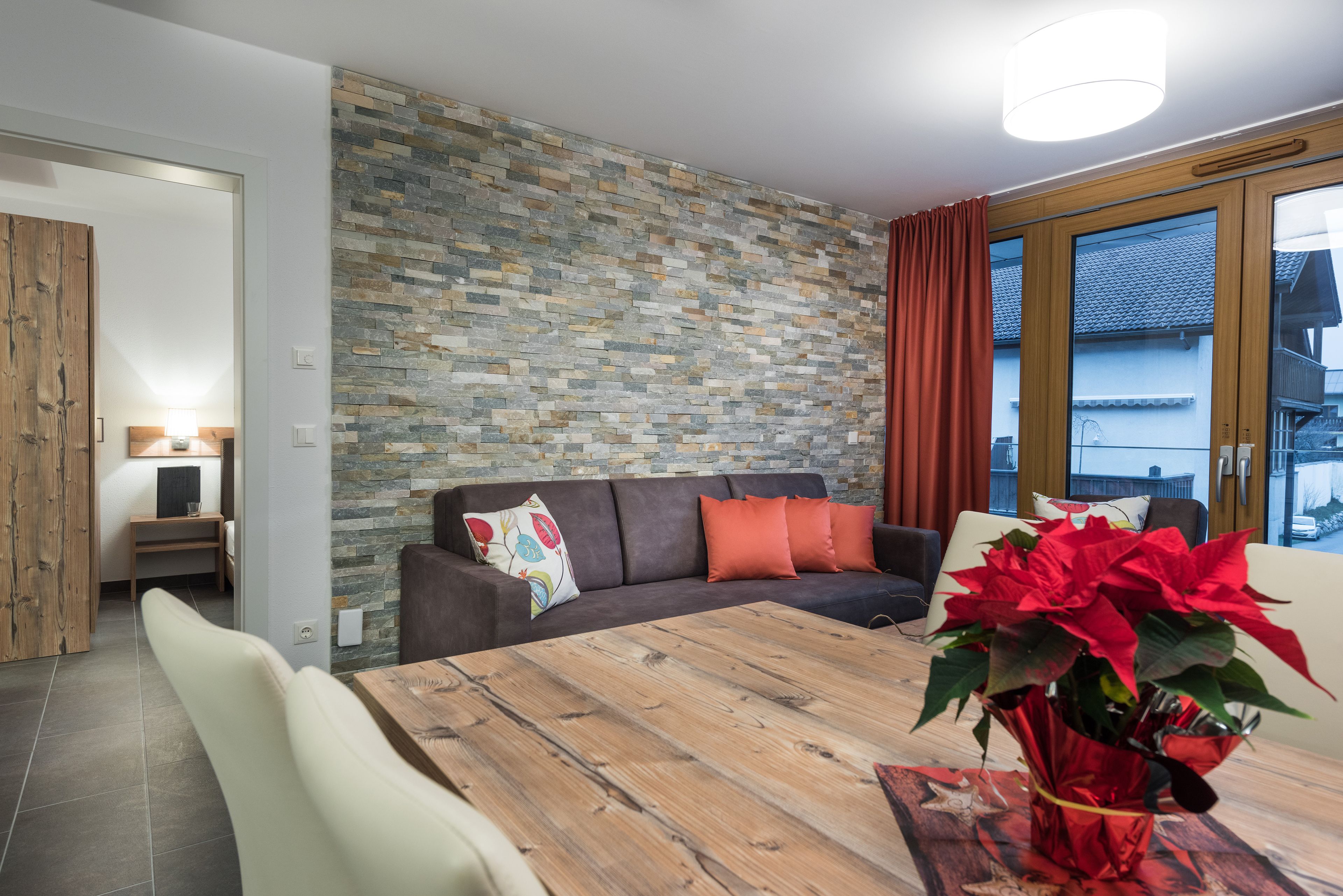 Una sala de estar moderna con una pared de piedra, una mesa de comedor de madera, un sofá con cojines naranjas y una planta en maceta roja.