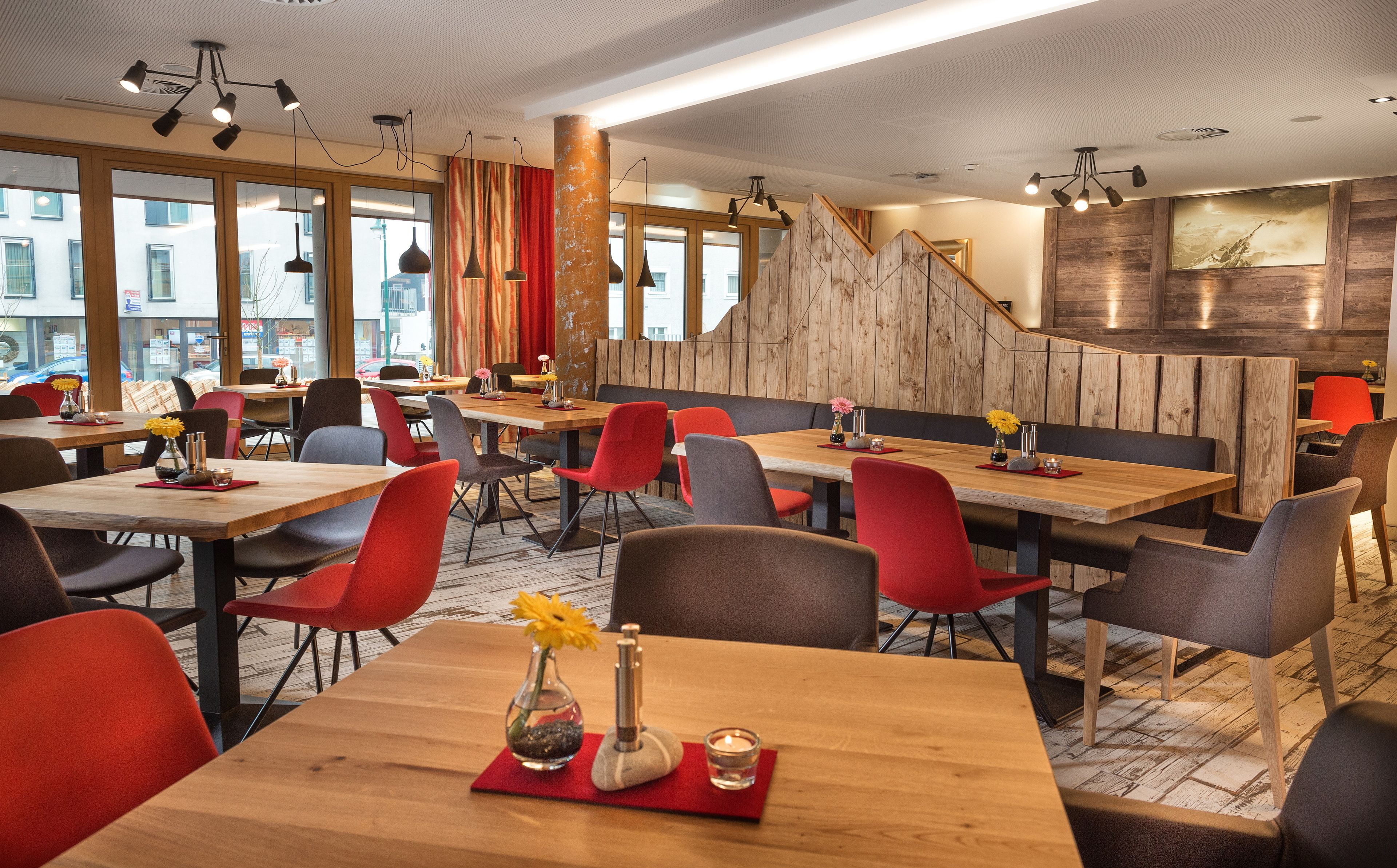 A modern restaurant interior with wooden tables, red and grey chairs, and a wooden accent wall.