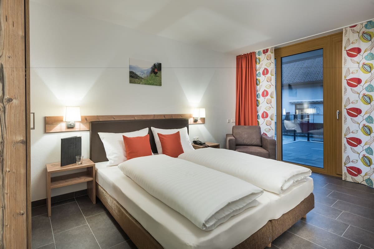 A hotel room with a large bed, a wooden headboard, a nightstand, a chair, and a view of the outside through a glass door.