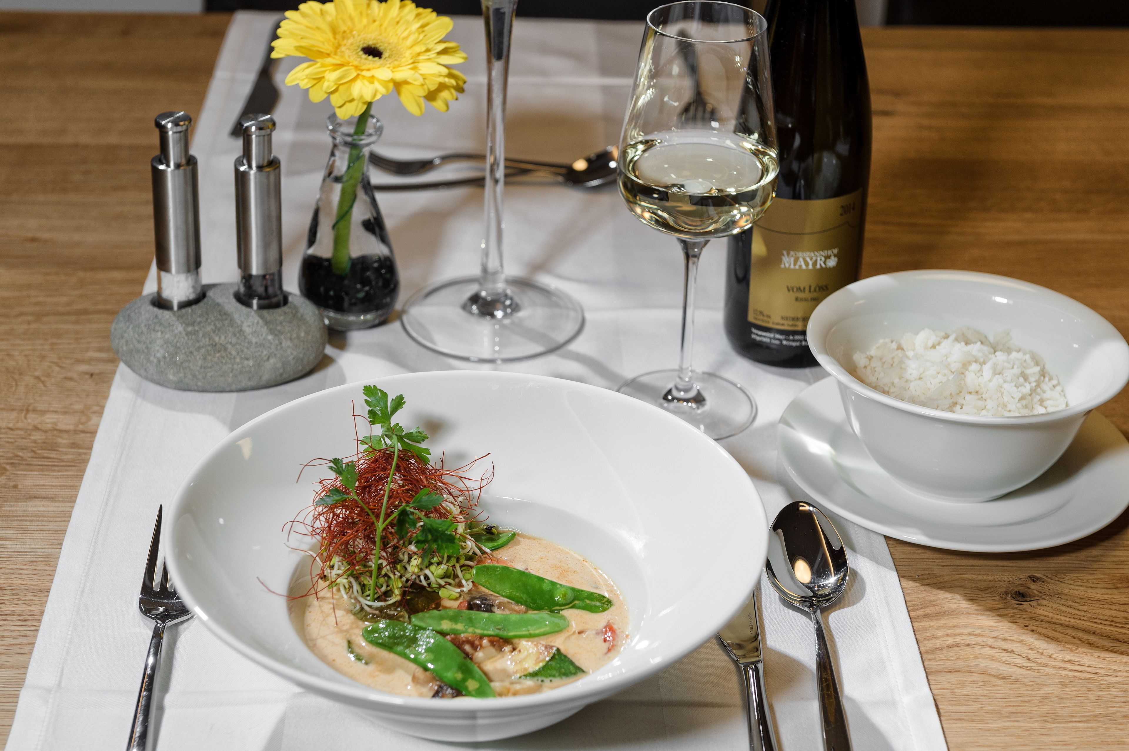 Una mesa servida con un plato de comida, una botella de vino, una copa de vino, un florero con una flor amarilla y saleros y pimenteros.