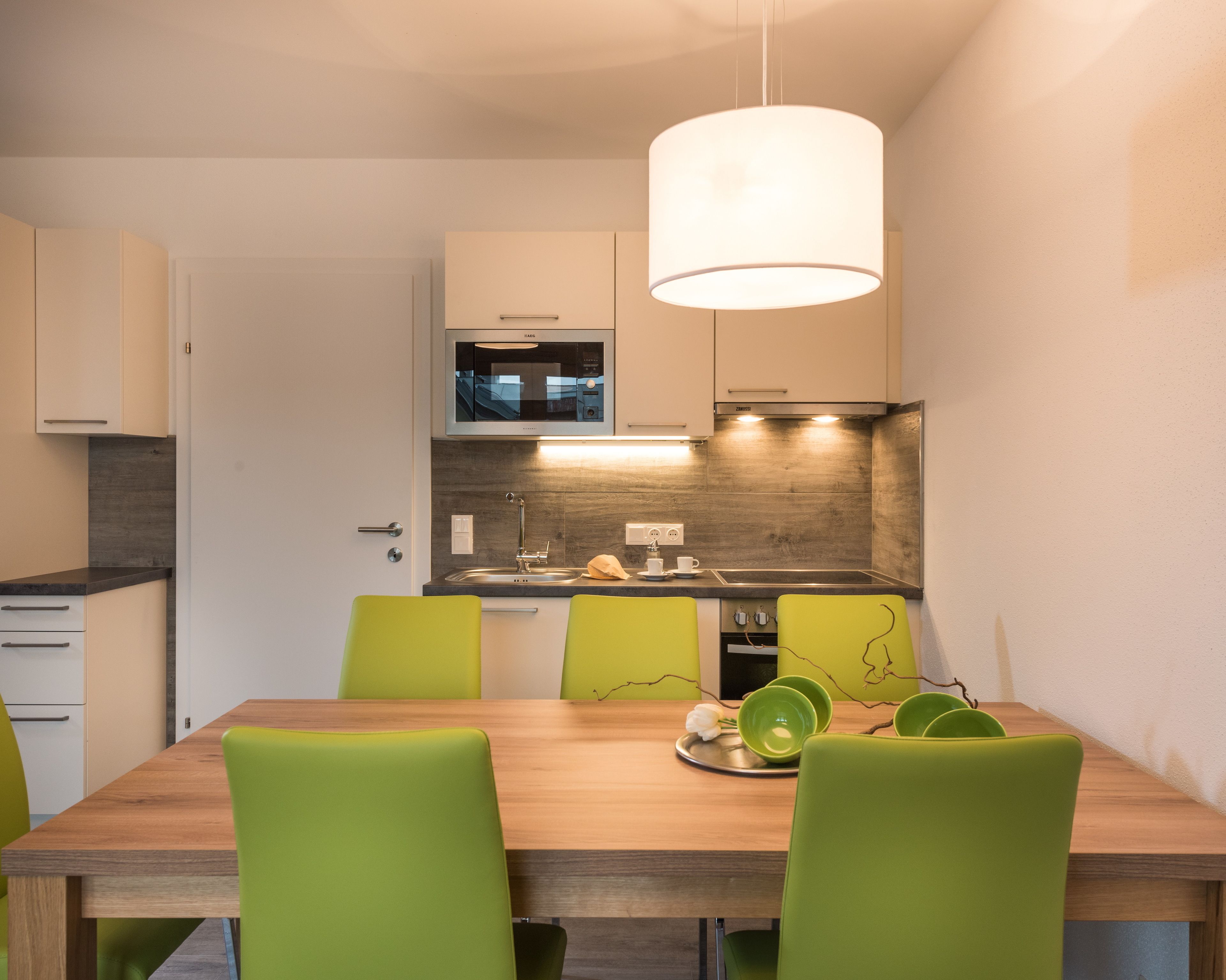 A modern kitchen with a wooden table and green chairs, a microwave, and a sink.