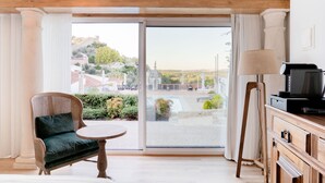 Una habitación con un amplio ventanal corredero que da a una zona de piscina, una silla de mimbre con cojín, una mesita redonda y un mueble de madera con un televisor.