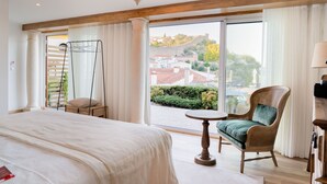 Habitación de hotel con una cama grande, una silla de mimbre, una mesita y vistas a un pueblo a través de amplios ventanales.