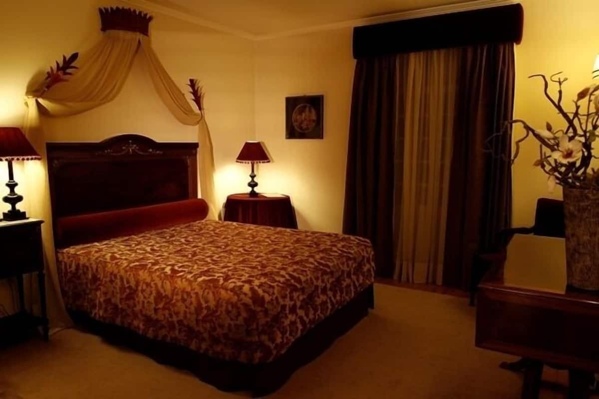 A hotel room with a large bed, two bedside tables with lamps, a nightstand, a vase with flowers, and a window with curtains.