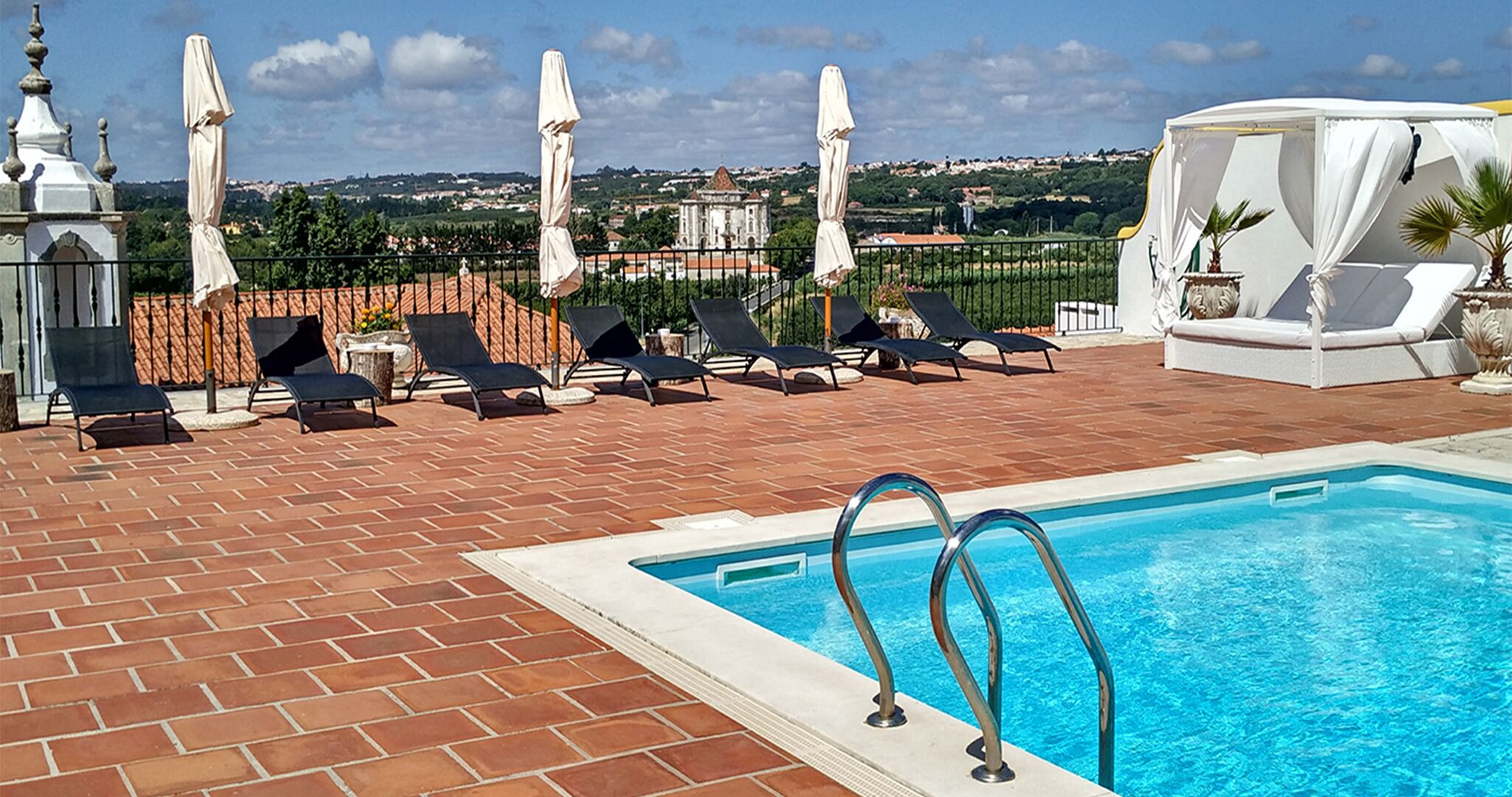 Un área junto a la piscina con sillones de descanso y sombrillas, con vista despejada a un edificio histórico en el fondo, y un patio de baldosas.