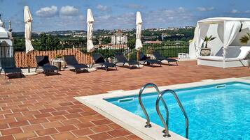 Un área junto a la piscina con sillones de descanso y sombrillas, con vista despejada a un edificio histórico en el fondo, y un patio de baldosas.