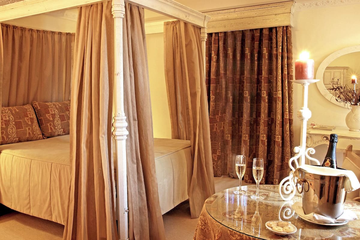 A four-poster bed with beige bedding, a bedside table with a lamp, and a table with two glasses and a bottle.