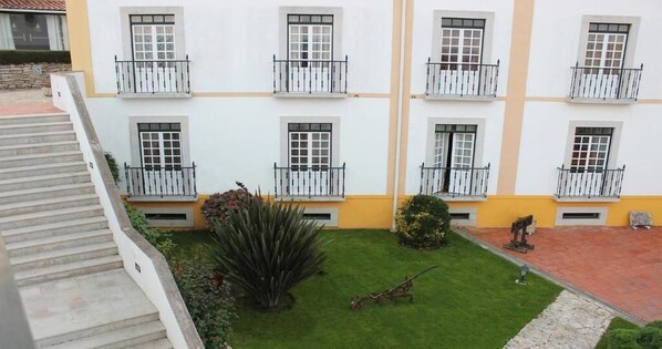 Un edificio de varios pisos con balcones, un pequeño jardín con césped y plantas, y un patio empedrado.