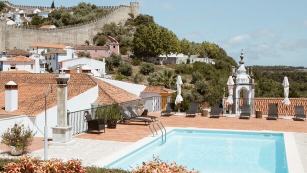Una piscina con tumbonas y sombrillas, rodeada de edificios y una colina con una estructura parecida a un castillo al fondo.