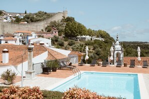 Una piscina con tumbonas y sombrillas, rodeada de edificios y una colina con una estructura parecida a un castillo al fondo.