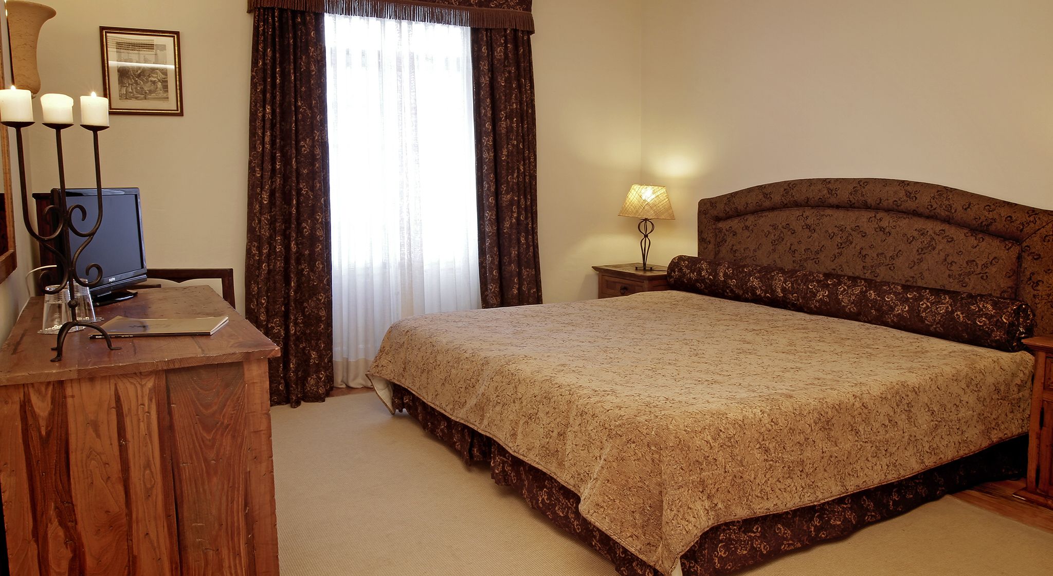 A bedroom with a bed, a wooden desk with a computer, a lamp, and a framed picture on the wall.