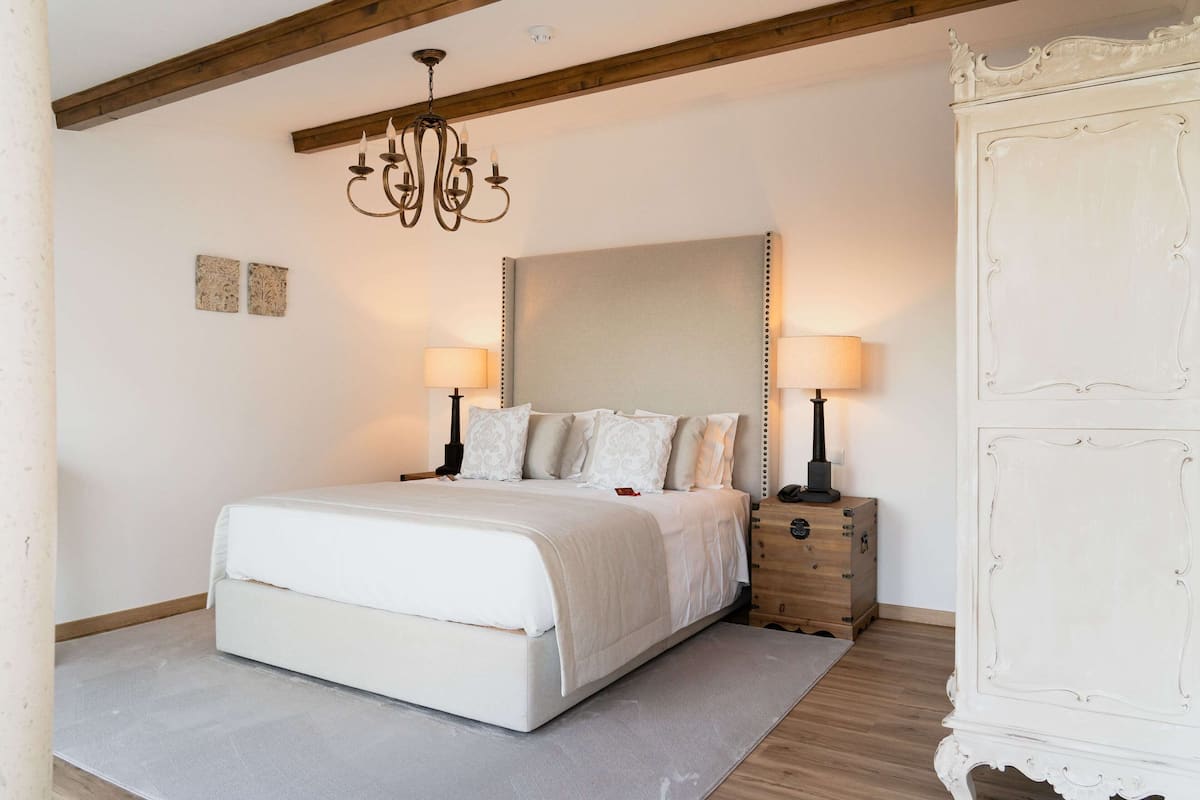A bedroom with a large bed, two bedside lamps, a wooden dresser, and a chandelier.