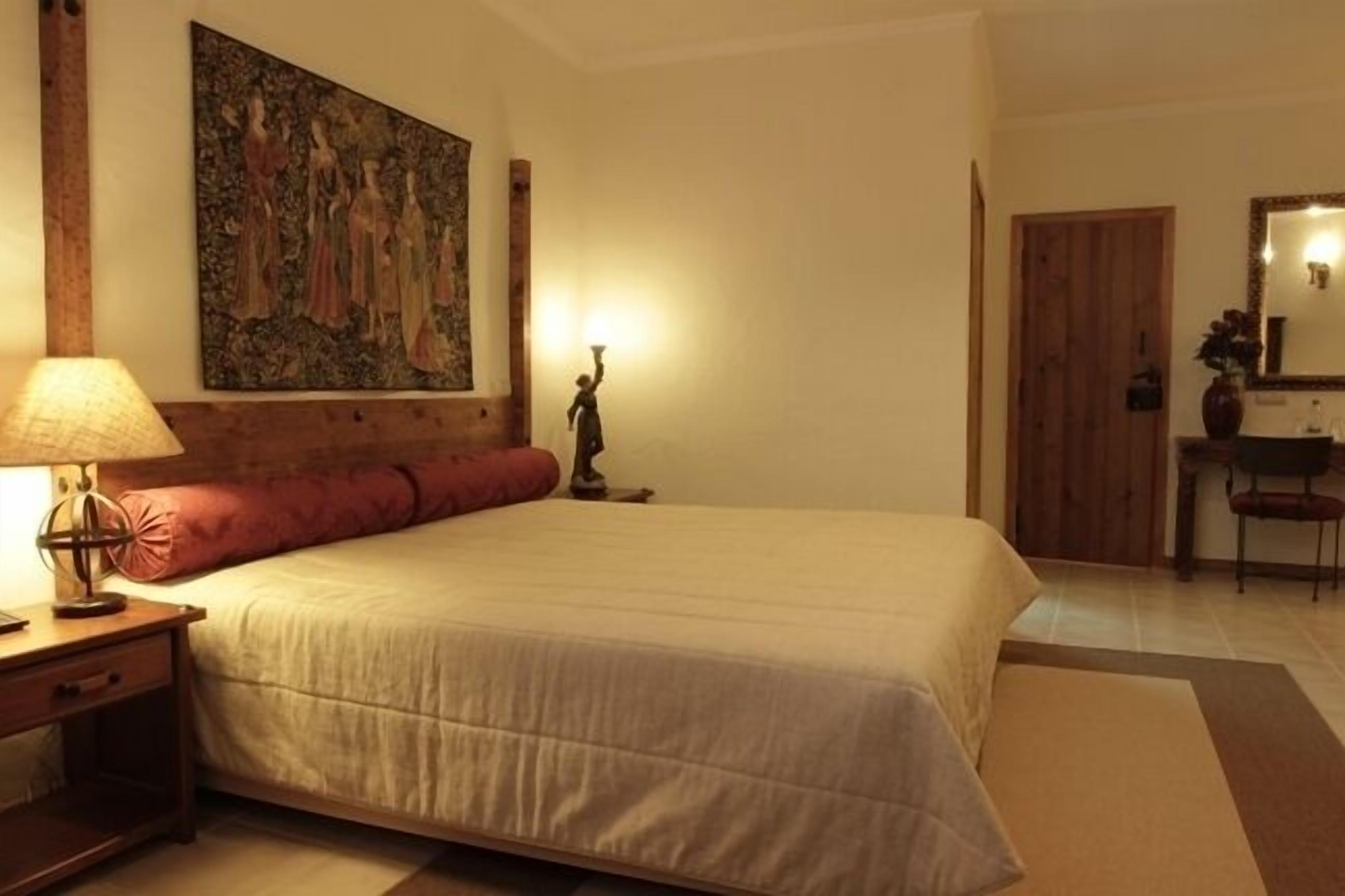 Un dormitorio con una cama grande, una mesita de noche con lámpara, un cuadro en la pared, una puerta de madera y un espejo.